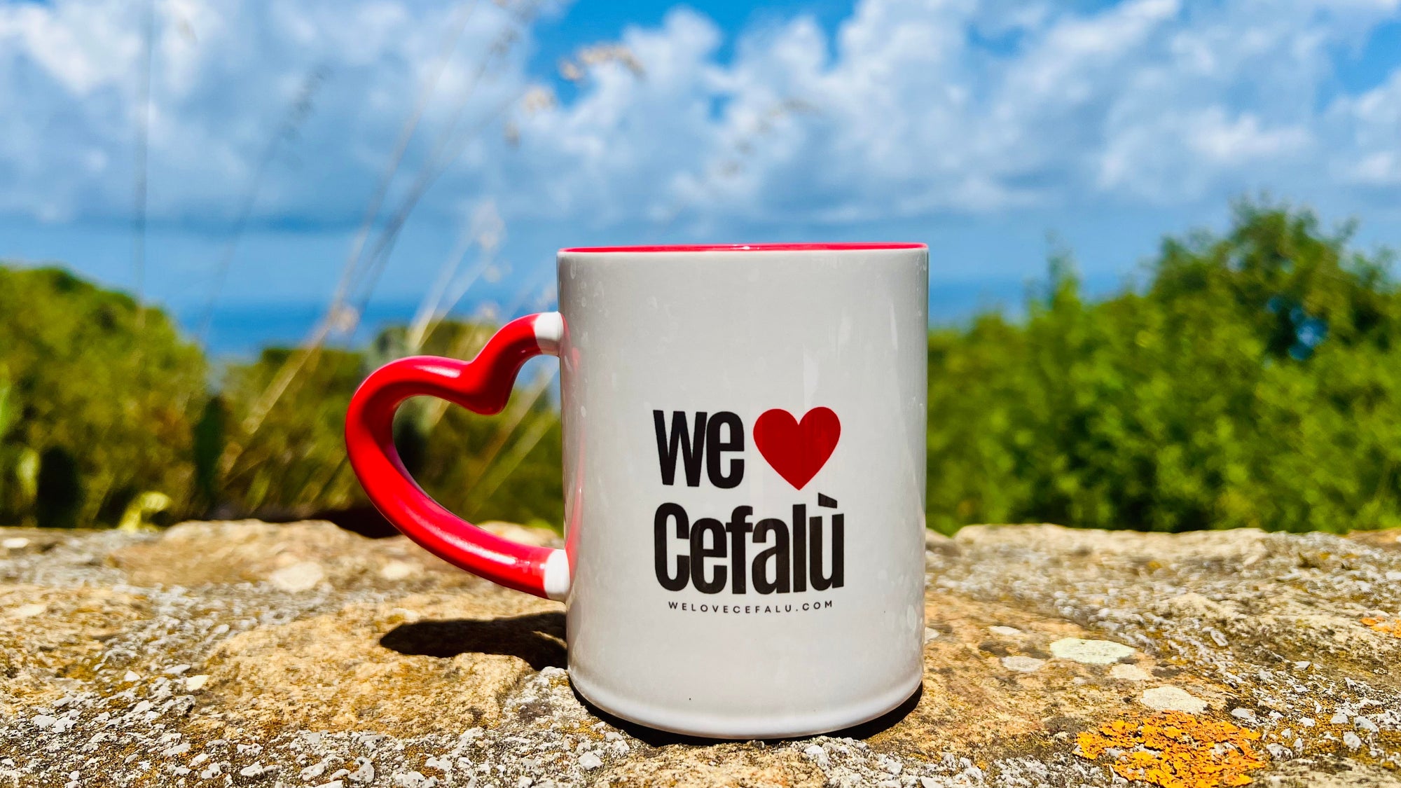 A white ceramic mug with a red handle and red interior sits on a stone surface outdoors, with a heart-shaped red graphic and the text 'we Cefalù' printed on the front. The background shows greenery and a blue sky with clouds.